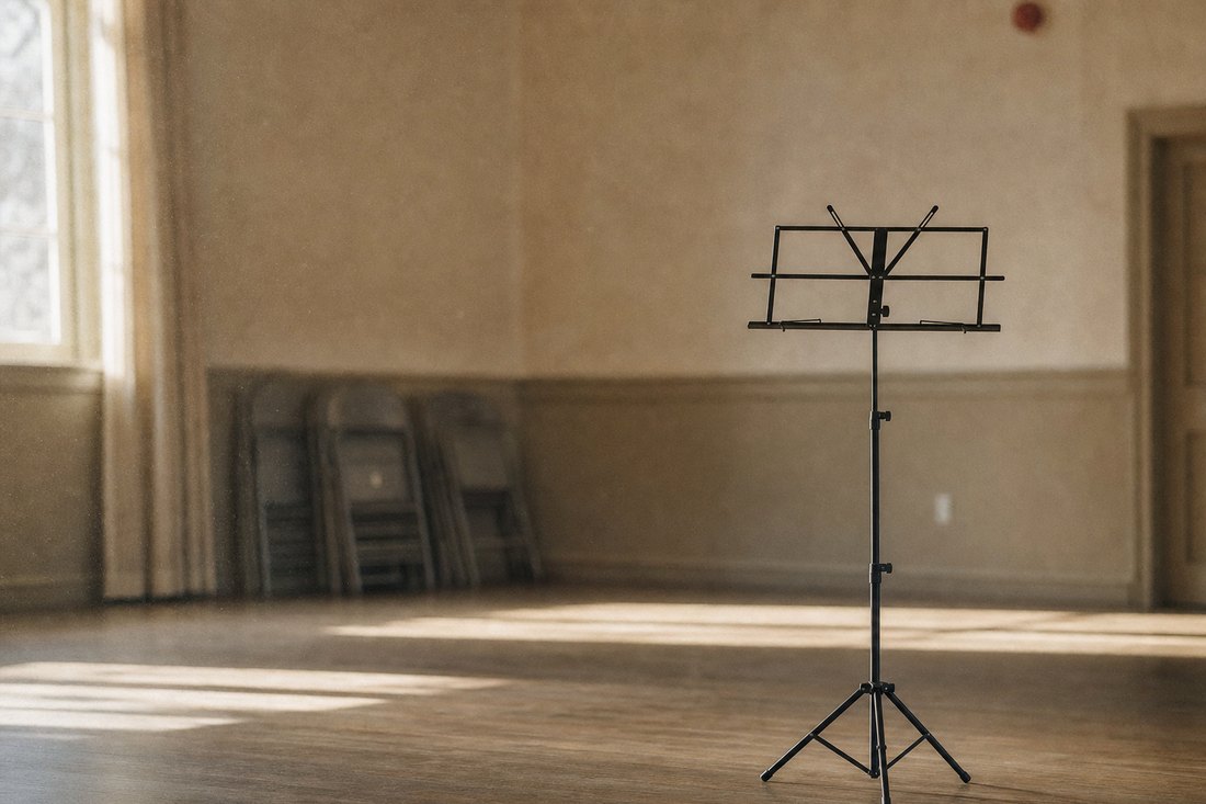 A single empty black folding music stand alone in a quiet community hall, late afternoon light raking across the floor, stacked folding chairs blurred behind it.
